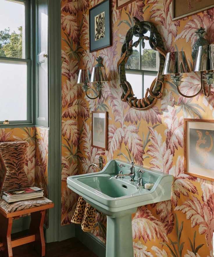 A small bathroom with orange and pink botanical wallpaper, mint green pedestal sink, and vintage wall decor