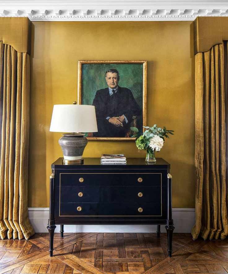 Black chest of drawers on a wooden floor, walls decorated in yellow with coving and yellow curtains with pelmets